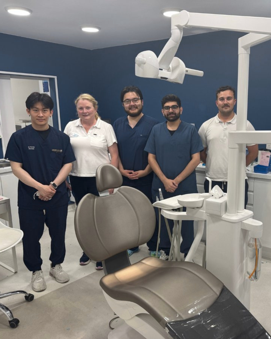 Dental team standing in a modern clinic wearing navy medical scrubs beside a dental chair and equipment.